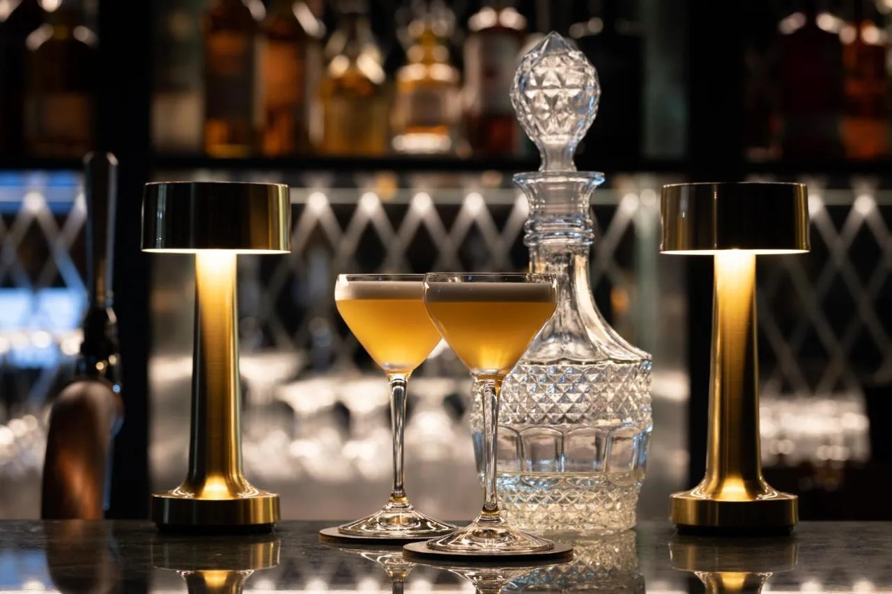 two glasses on a table with a bottle and two lights at Sun Street Hotel Shoreditch in London